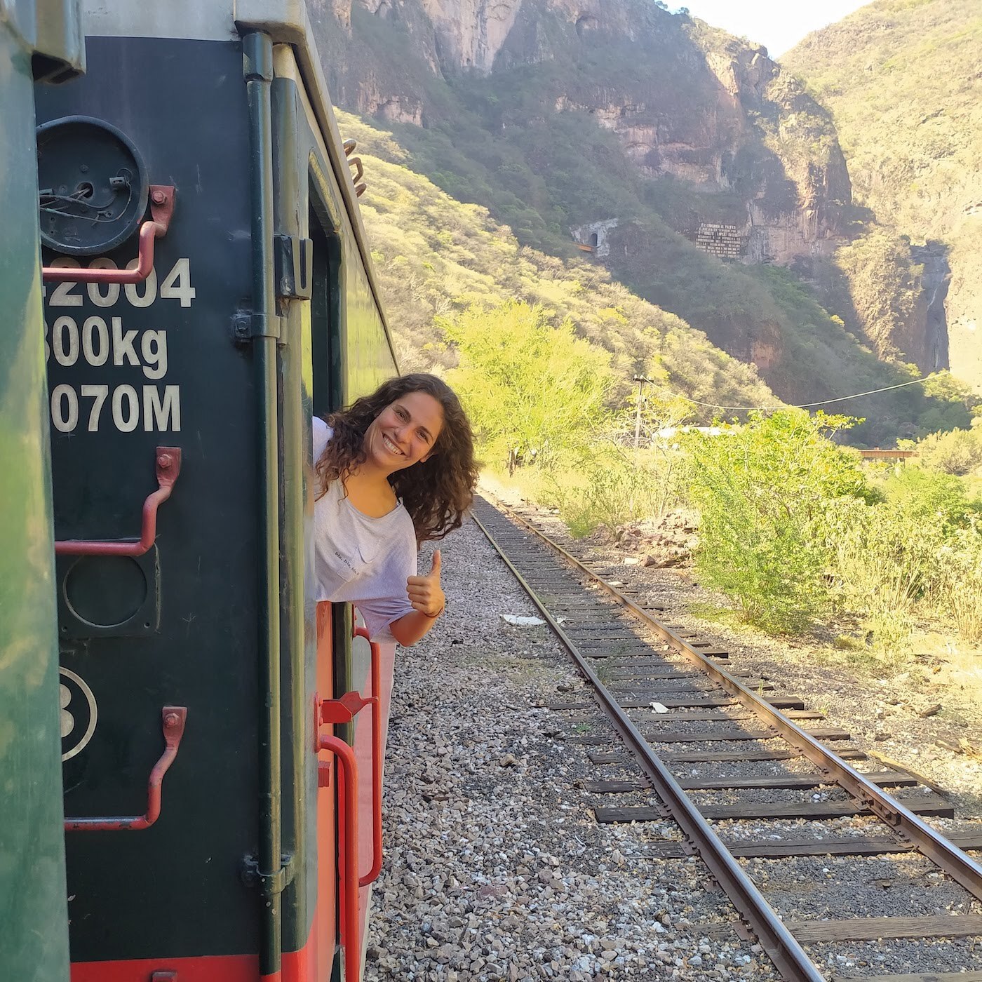 Una persona sonriente asomada por la ventana de un tren, con un pulgar hacia arriba, rodeada de un paisaje montañoso y vegetación cercana.