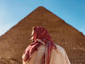 Hombre de espaldas con una vestimenta tradicional árabe, frente a la Gran Pirámide de Giza bajo un cielo despejado.